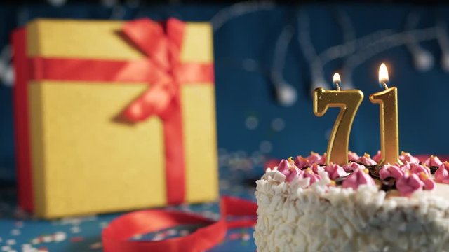 White birthday cake number 71 golden candles burning by lighter, blue background with lights and gift yellow box tied up with red ribbon. Close-up