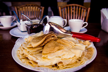 Fried pancakes are laid out on a plate with a slide, next to a shovel, cups, jam, sour cream, chairs. Maslenitsa. Dark background. Russian food