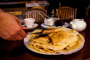 Female hand takes a pancake from a plate. Pancakes are laid out on a plate with a slide, next to a shovel, cups, jam, sour crea m, chairs. Maslenitsa. Dark background. Russian food