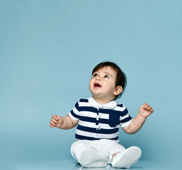 Little child in striped t-shirt, white pants and booties. He is looking up, sitting on floor against blue background. Close up