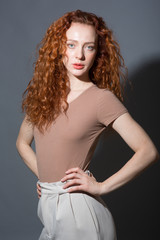 Serious red-haired curly-haired woman, looking directly at the camera, holding her hands on her belt. On an isolated gray background