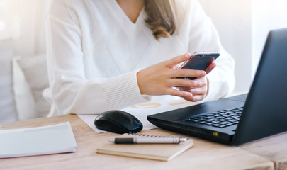 Close up of hand young female using mobile phone to searching information while she working with laptop on desk