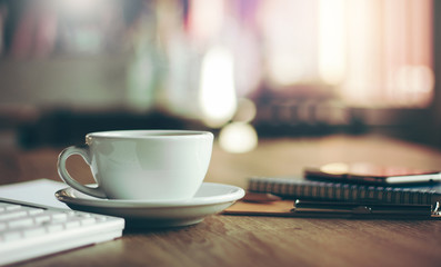 Closeup white cup of coffee with smartphone, notebook  on wooden table in cafe. with vintage light, blurred and bokeh background