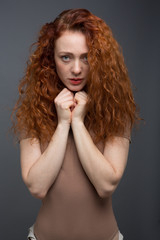 A serious red-haired curly-haired woman, holding her hand near her chin, thinking about something. On an isolated gray background