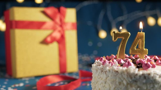 White birthday cake number 74 golden candles burning by lighter, blue background with lights and gift yellow box tied up with red ribbon. Close-up