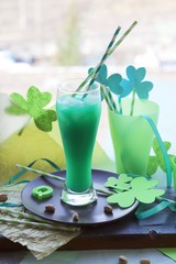 St. Patrick Day, glasses with a green drink, cocktail tubes with a decorative shamrock, clover on a wooden surface on a window background, getting ready for a party