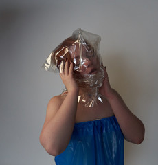 Portrait of a young redhead girl on grey background with a plastic bag on her head. The concept of plastic pollution of nature. Excess plastic in a person's life
