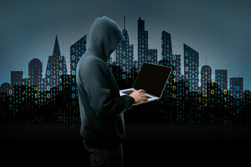 unknown hacker in the hood with laptop over background of city silhouette with binary code
