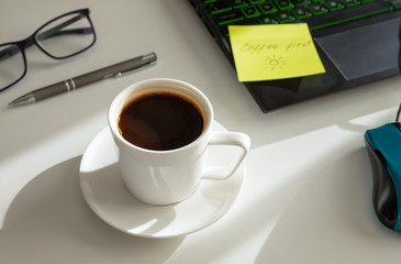 A cup of coffee in the workplace, a pen, a laptop, glasses. Hard light. Coffee first concept.