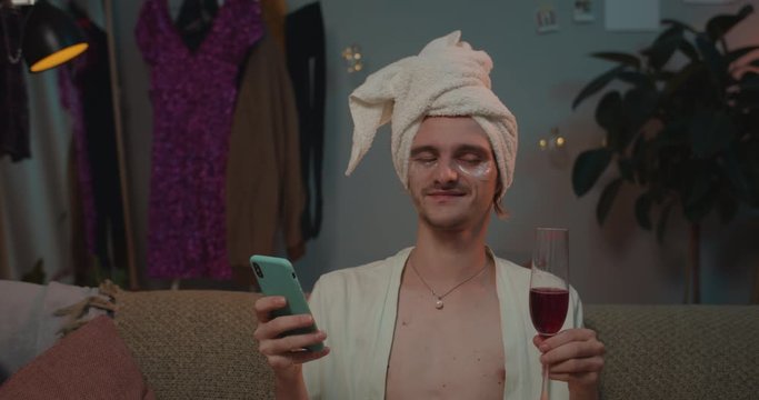 Close up of relaxed trans man in home wear using smartphone and drinking wine. Young transgender enjoing free time while sitting on sofa and surfing net. Home background.
