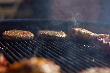 A portrait of the smoke in the middle of a hot barbecue grill. With the meat resting at the side of the bbq. There are hamburgers, chicken drumsticks and spareribs on the steel grill.