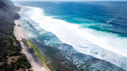 A done shot of Nyang Nyang Beach, Bali, Indonesia. The waves are rushing to the shore, making the water bubbly. The beach is covered with green algae, further on it's sandy. Tall cliffs on the side