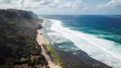 A done shot of Nyang Nyang Beach, Bali, Indonesia. The waves are rushing to the shore, making the water bubbly. The beach is covered with green algae, further on it's sandy. Tall cliffs on the side