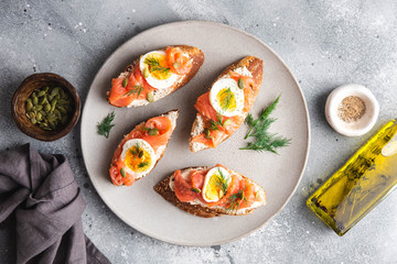 Italian appetizer bruschetta with red fish, boiled egg, cream cheese and dill on a grey plate on a grey background, close up horizontal