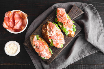 Italian appetizer bruschetta with parma ham, arugula and grated parmesan on a dark wooden board,...