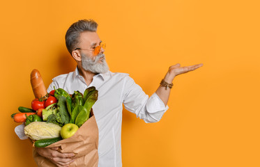 Cool old mature senior man with gray beard shopping hold grocery shopping bag with healthy organic vegetables on yellow - Powered by Adobe