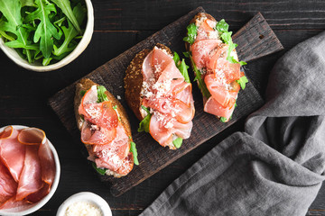 Italian appetizer bruschetta with parma ham, arugula and grated parmesan on a dark wooden board,...