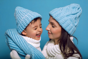 mother and son in white sweaters and blue hats stand on a blue background