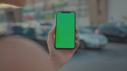 Lviv, Ukraine - May 19, 2018: Close-up of young urban girl showing vertical smartphone with greenscreen chrome key technology on parking lot outside. City life and modern gadgets.