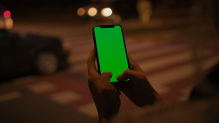 Lviv, Ukraine - May 19, 2018: Young woman hitchhiking on night highway holding and using modern mock-up vertical smartphone greenscreen outdoors. Close-up.
