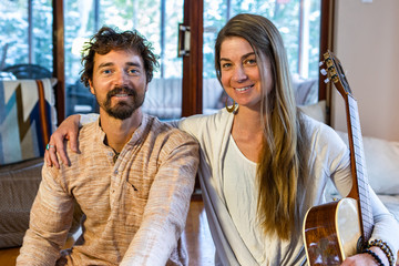 Happy shamanic man and woman in room holding musical instruments like classical guitar looking at camera while preparing for calm peaceful meditation