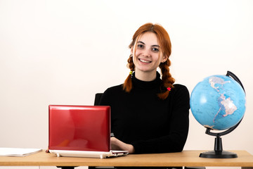 Happy young office worker woman sitting behind working desk with laptop computer, cell phone, notebook and geographic globe of the world.