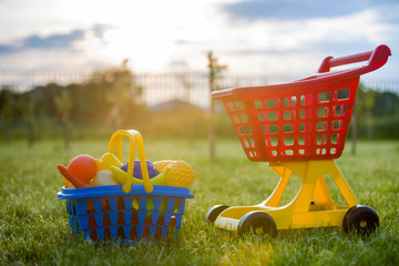 Shopping pushcart and a basket with toy fruits and vegetables. Bright plastic colorful toys for children outdoors on sunny summer day.