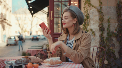 Happy young woman with red lipstick hat wearing stylish coat typing on phone during sunny day sitting in cafe on breakfast outdoors smile city fashion online stylish slow motion