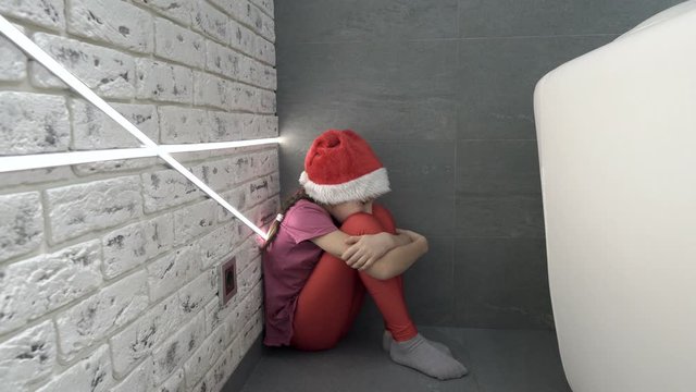 A little offended girl in a Santa Claus hat is sitting on the floor of the house in the corner, hugging her knees and wiping away tears. Closeup. Concept. 4K.