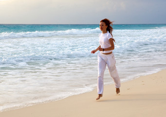 slender woman in a white tracksuit jogging along the edge of the waves on the sandy beach of the sea