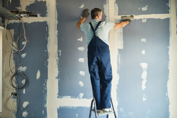 Full length back view of senior construction worker smoothing dry wall standing on ladder while renovating house, copy space