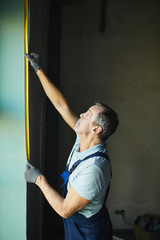Fototapeta na wymiar Side view of senior construction worker measuring wall with tape while renovating house, copy space