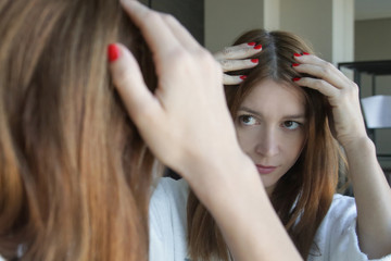 Fototapeta premium Portrait of a beautiful young woman examining her scalp and hair in front of the mirror, hair roots, color, grey hair, hair loss or dry scalp problem