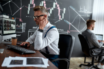 Satisfying the needs. Stylish businessman, trader sitting in front of computer monitor while working using wireless earphones. His colleague and blackboard full of data analyses in the background.