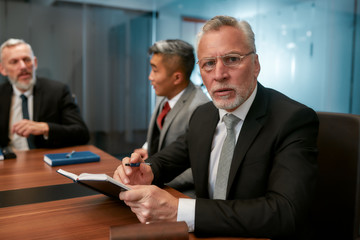 Portrait of handsome mature man in formal wear and eyeglasses looking at camera while sitting at the office table with his coworkers.