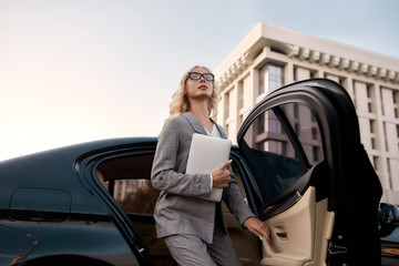 Female leader. Young beautiful business woman in stylish suit holding laptop and getting out of a black car. Business concept