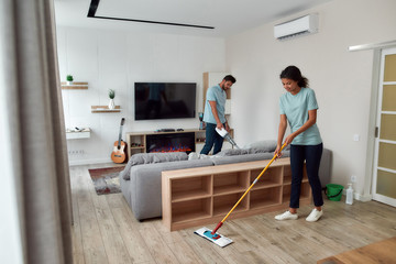 Cleaning team. Two professional cleaners in uniform working together in the living room. Young afro american woman cleaning floor with mop while her male coworker cleaning sofa with handheld vacuum