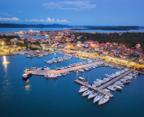 Fototapeta na wymiar Aerial view of boats and yachts in port and city at night. Summer landscape with city lights, buildings, illuminated streets, mountain, motorboats, blue sea, sky at dusk. Top view. Travel in Croatia