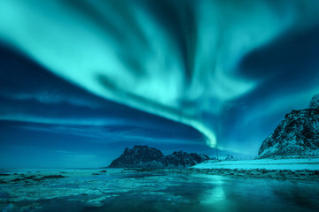 Aurora borealis über den schneebedeckten Bergen und dem Sandstrand im Winter. Nordlichter auf den Lofoten, Norwegen. Sternenhimmel mit Polarlichtern. Nachtlandschaft mit Aurora, gefrorener Meeresküste, blauem Himmel