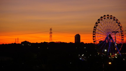 Naklejka premium Ferris wheel at beautiful sunset. Red or orange sky background. Big ferris wheel at amusement park in orange sunset. Silhouette giant swing on twilight time of the day. Copy space. Textured wallpaper.