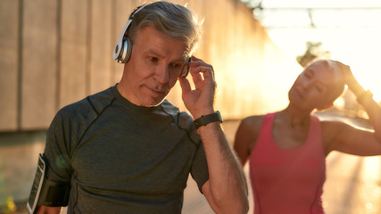 Morning workout. Handsome and fit middle-aged man in sports clothing adjusting headphones while his wife doing stretching exercises