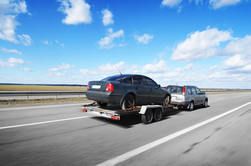 A crossover with trailer towing a car on the countryside road in motion against sky