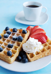 Fresh Viennese or Belgian waffles on a porcelain plate with fresh strawberries and blueberries and whipped cream and a cup of black coffee nearby on a pastel blue background. Dessert for breakfast.