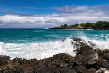 Wave breaking on rough shore of the Kona coast, on Hawaii's Big Island. Blue-green Pacific ocean beyond; rocky shoreline with trees in the distance. Cloudy blue sky above.