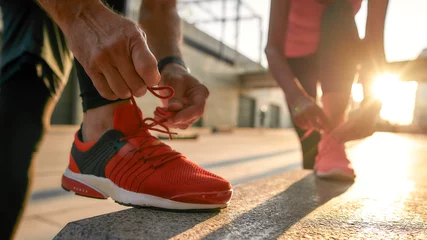 Fotobehang Exercising together. Close up photo of two people in sport clothes tying shoelaces before jogging outdoors. Fit, fitness, exercise © Svitlana