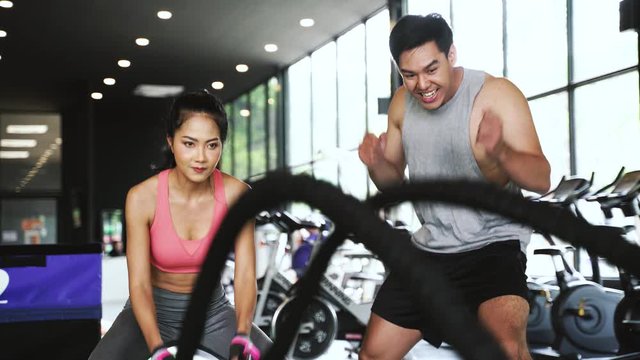 Asian personal trainer coaching a bodybuilding woman to perform crossfit battle ropes exercise in the fitness gym. Workout training coach business concept