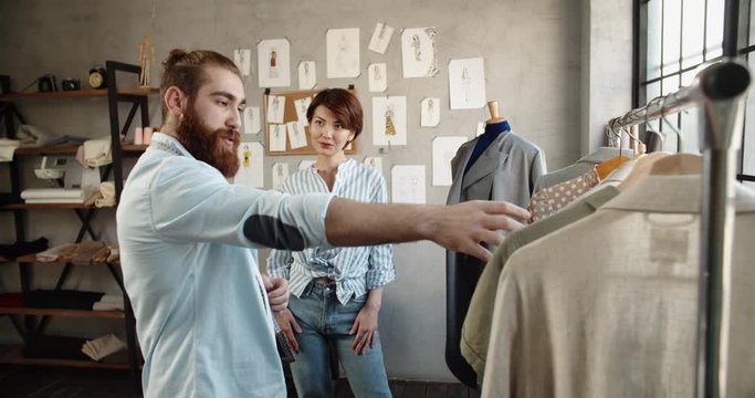 A happy multiethnic couple is working as tailors together, designing new clothes at their office, picking the best options for customers - small family business concept 4k footage