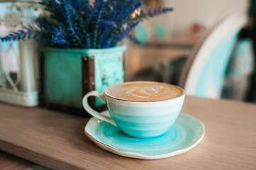 Cup of freshly brewed coffee with milk foam and a pattern in a turquoise-blue porcelain mug on a saucer against a background of violet flowers in a vase. Grab a coffee in the light interior of cafe.