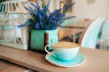 Cup of freshly brewed coffee with milk foam and a pattern in a turquoise-blue porcelain mug on a saucer against a background of violet flowers in a vase. Grab a coffee in the light interior of cafe.