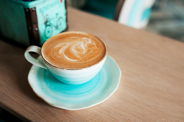 Cup of freshly brewed coffee with milk foam and a pattern in a turquoise-blue elegant porcelain mug. Grab a coffee in the beautiful light blue interior of the cafe.